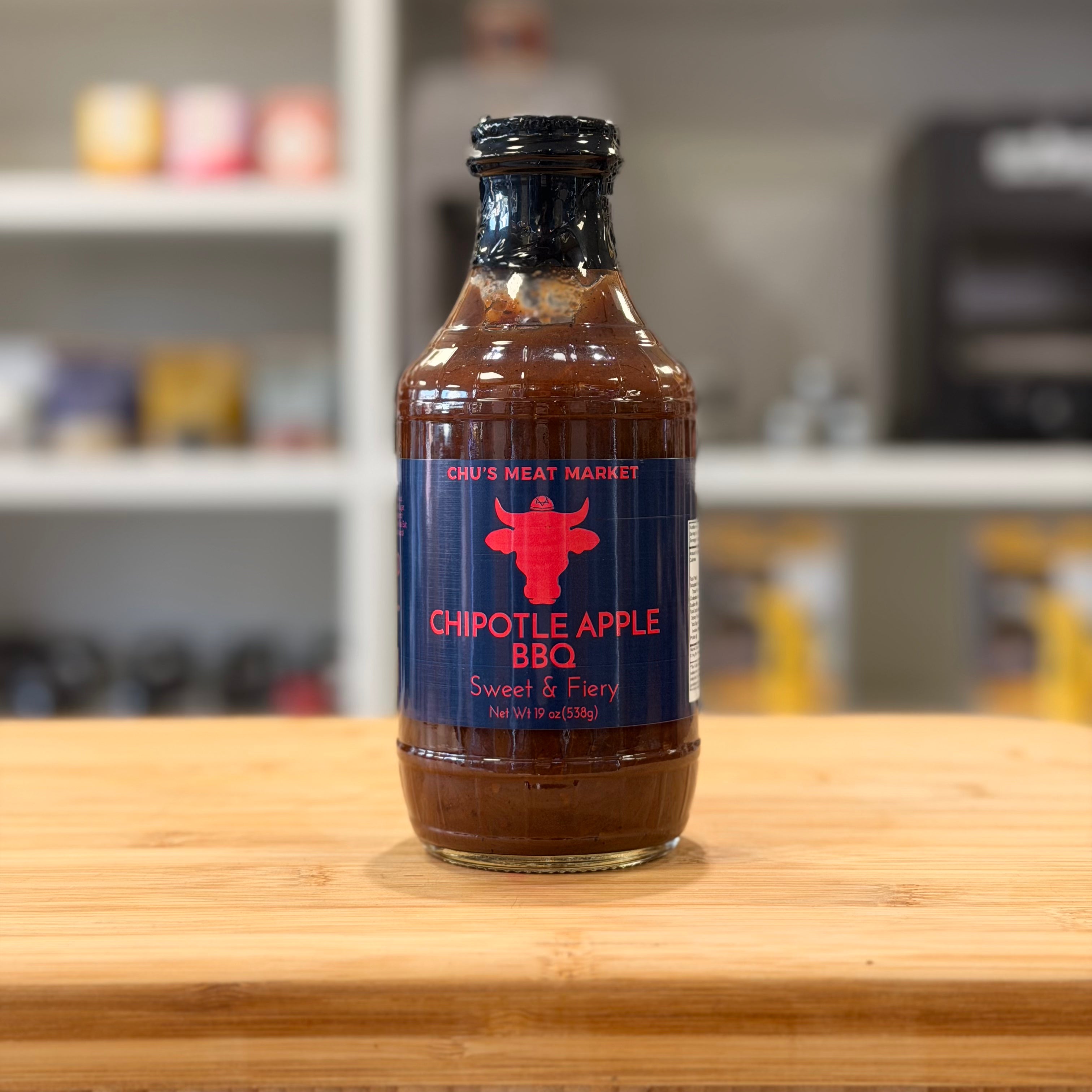 Bottle of Chu's Meat Market Chipotle Apple BBQ sauce on a wooden surface with a blurred background.