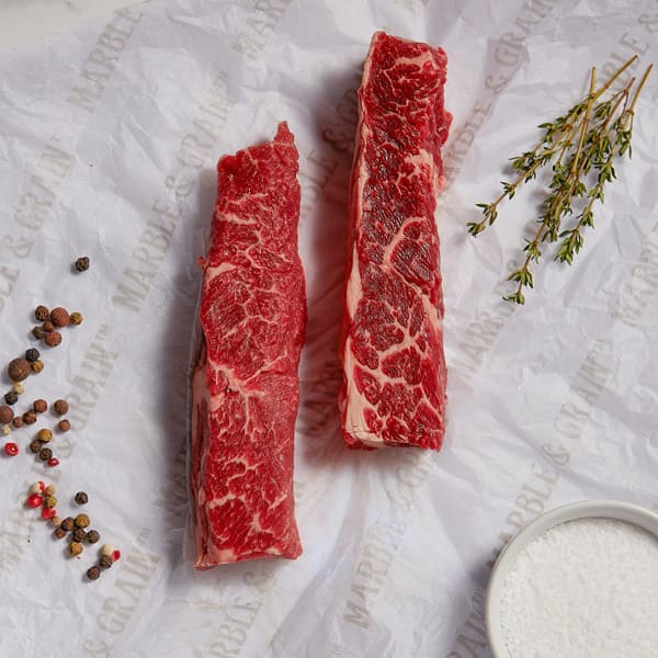 Two raw steaks on a textured white surface with spices and thyme.