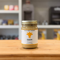 Jar of Dijon mustard from Chu's Meat Market on a wooden surface with a blurred kitchen background.