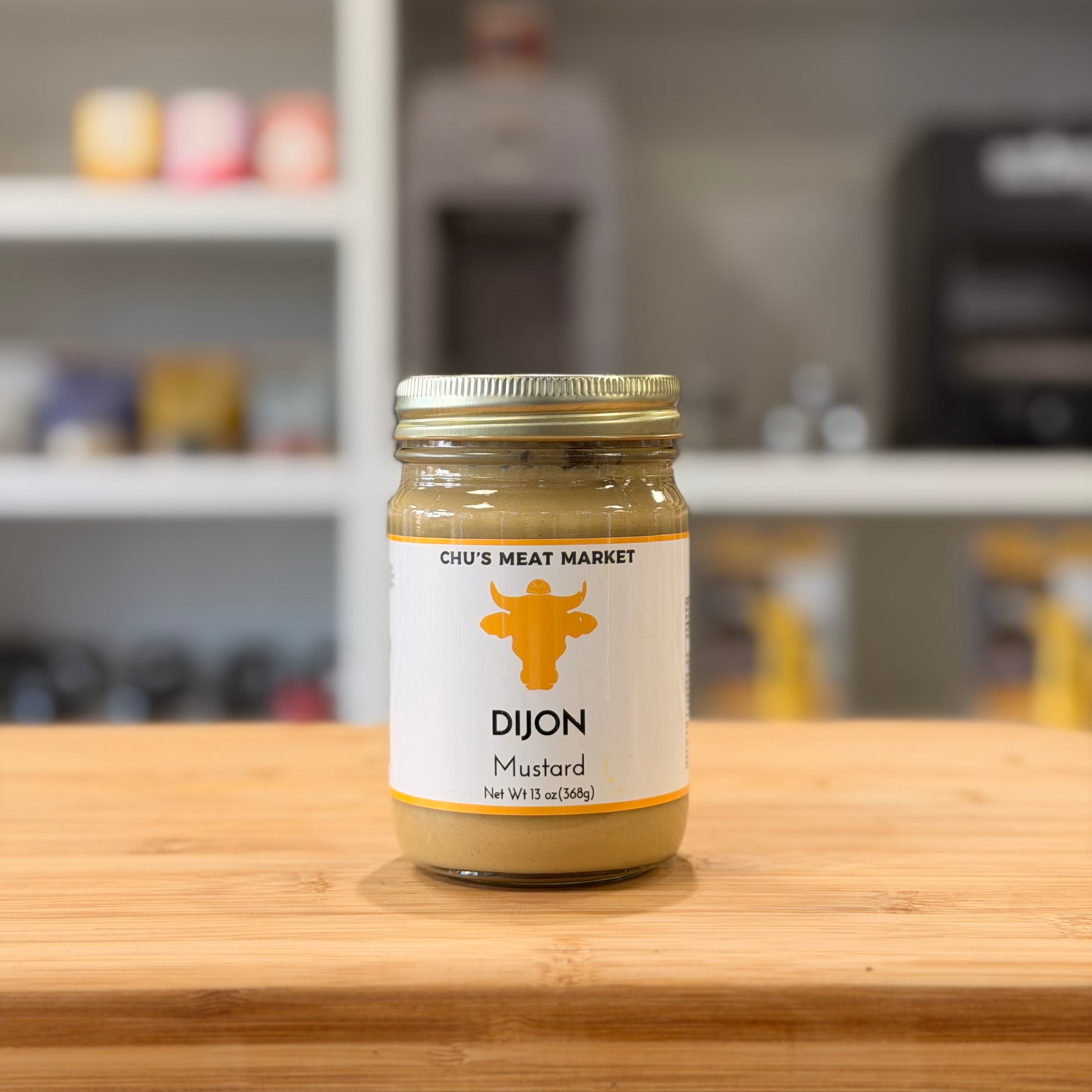 Jar of Dijon mustard from Chu's Meat Market on a wooden surface with a blurred kitchen background.