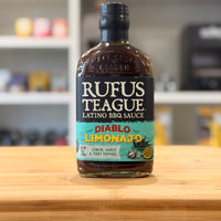 Bottle of Rufus Teague Latino BBQ Sauce Diablo Limonajo on a wooden surface with a blurred kitchen background.