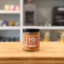 Jar of Hb Smoky Honey Habanero spice on a wooden surface with a blurred kitchen background
