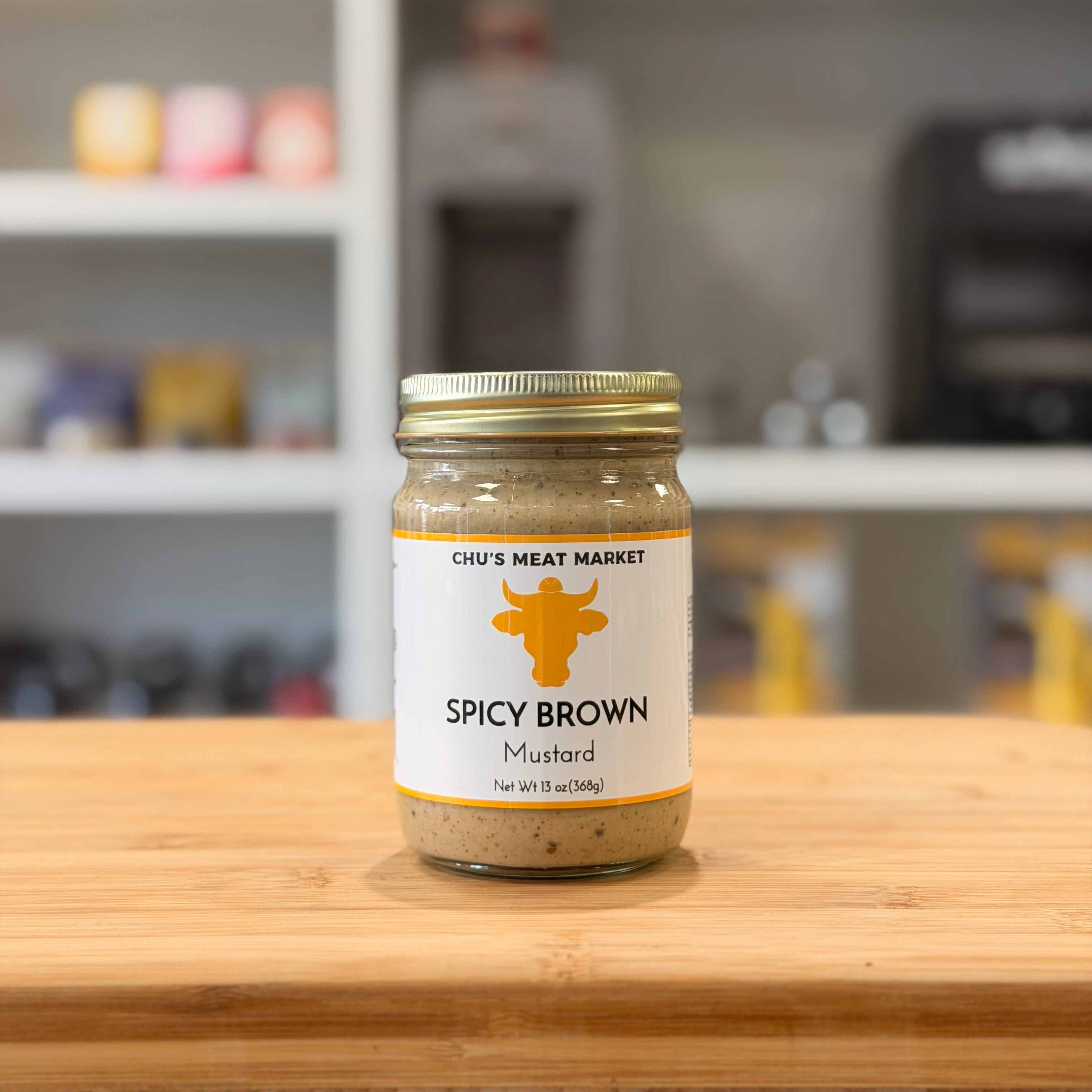 Jar of spicy brown mustard from Chu's Meat Market on a wooden surface with a blurred kitchen background.